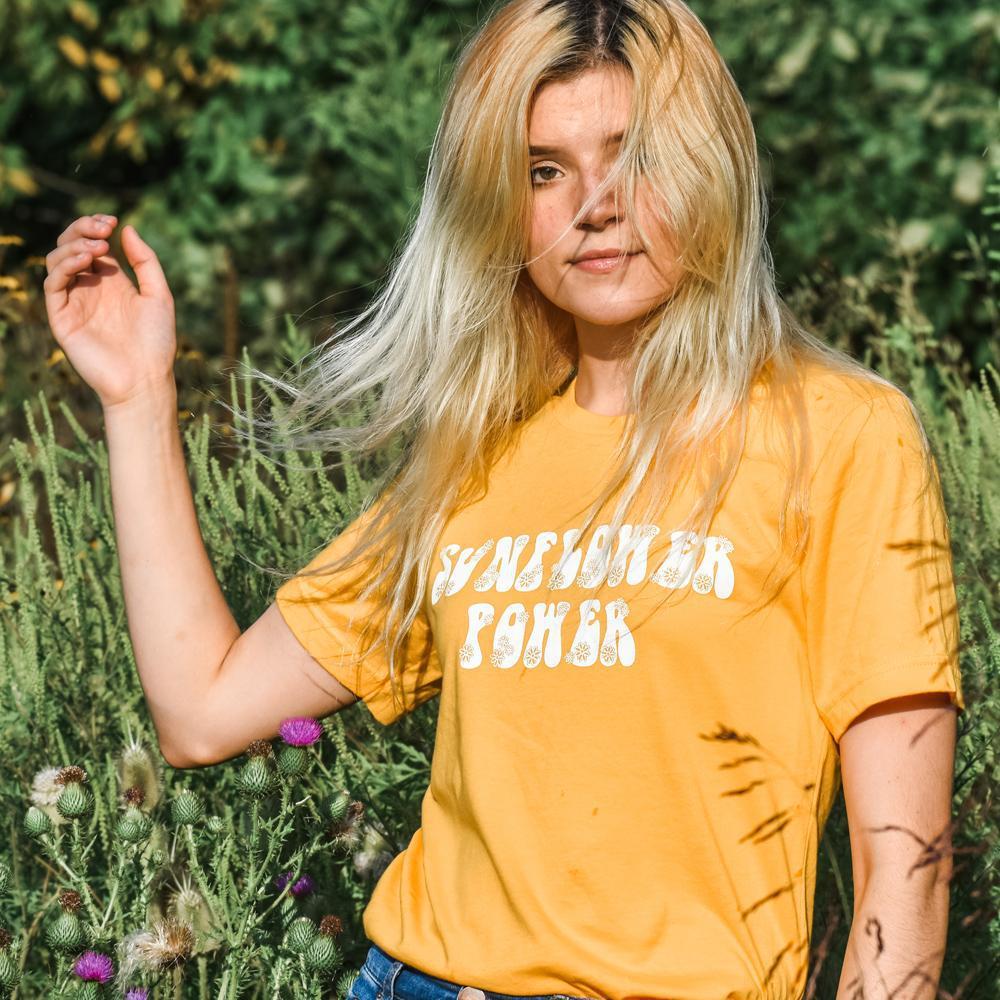 Woman wearing a yellow ‘Sunflower Power’ graphic tee with retro white flower lettering, standing in a sunny field