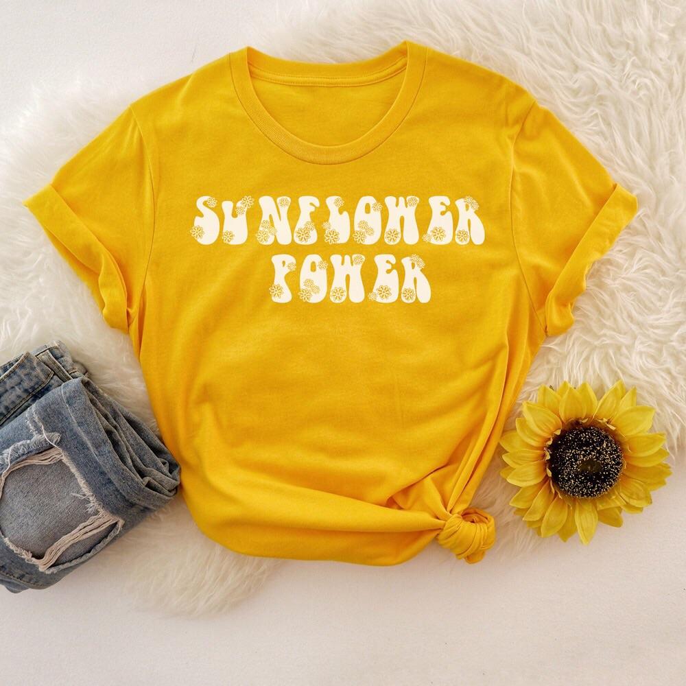 Yellow ‘Sunflower Power’ graphic tee with white retro bubble letters and floral accents, styled with denim jeans and a sunflower on a soft white background.
