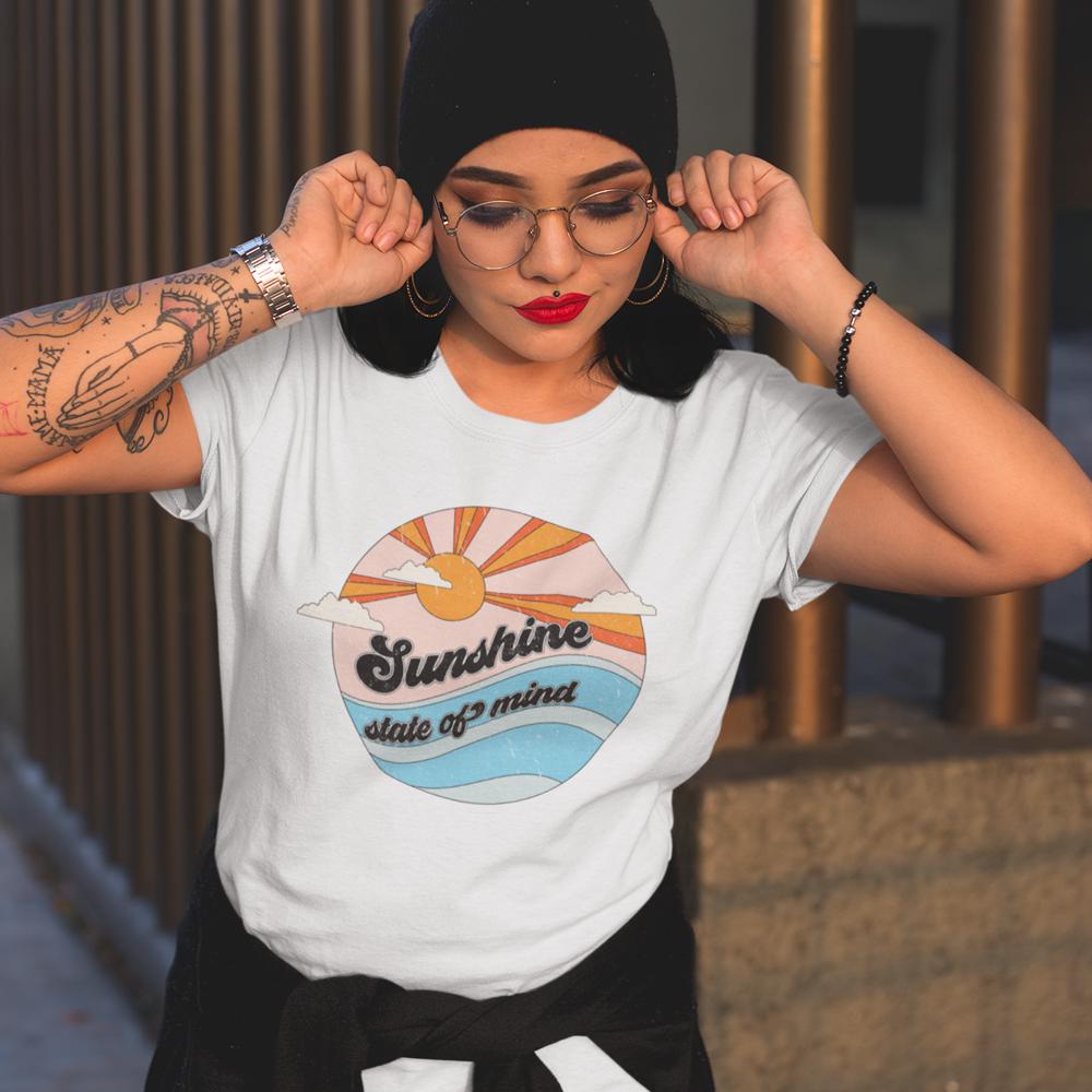 Retro-inspired white graphic t-shirt with ‘Sunshine State of Mind’ design featuring a sunrise and waves, styled with glasses and beanie, by Funnel Cake Tree.