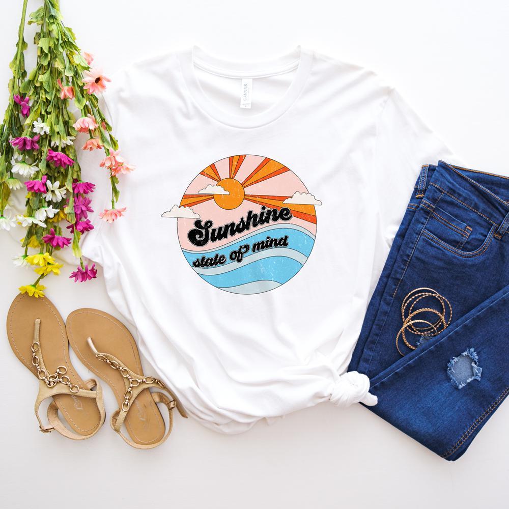 Flat lay of white t-shirt with retro ‘Sunshine State of Mind’ graphic featuring sun and waves, styled with denim jeans, sandals, and colorful flowers.