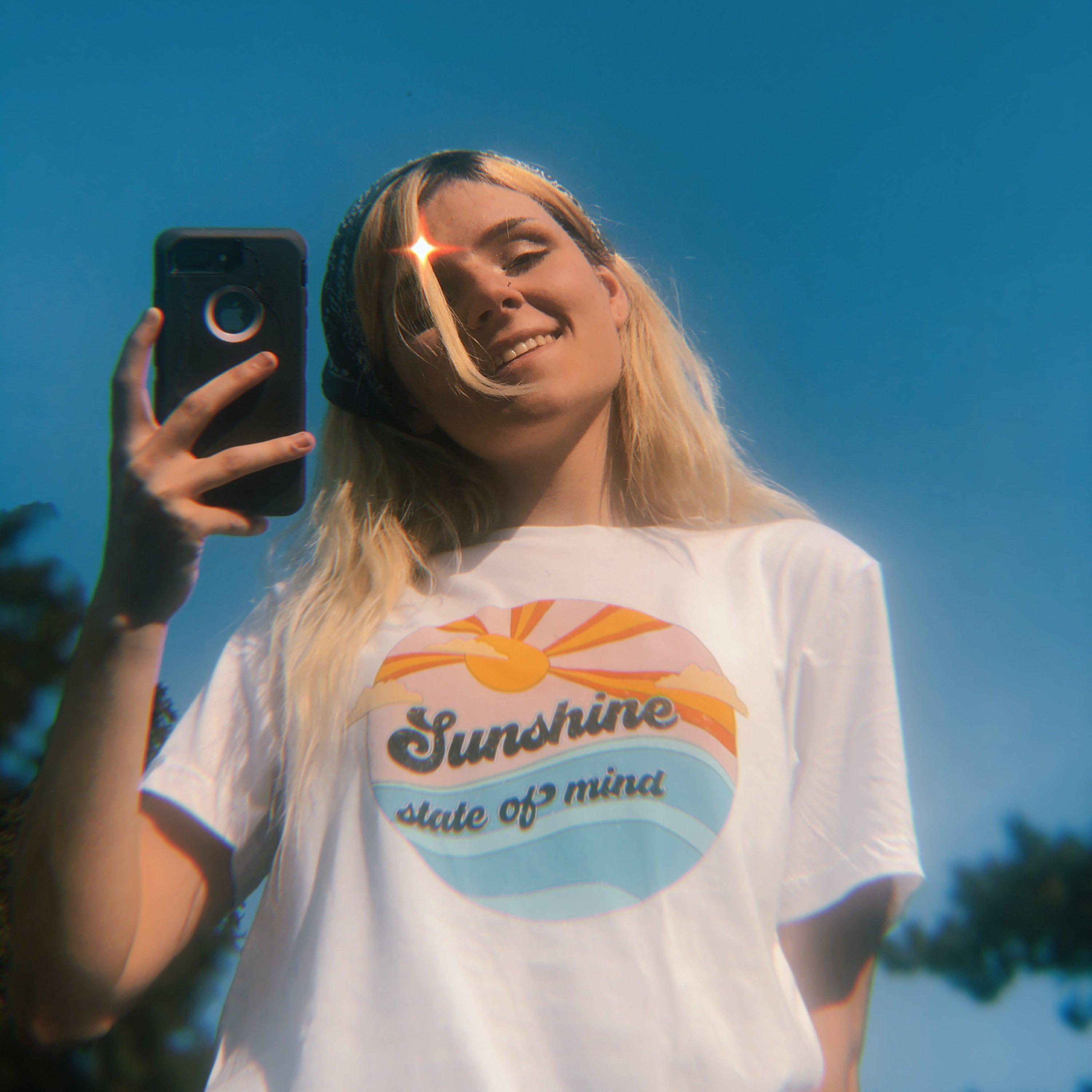 White t-shirt with retro ‘Sunshine State of Mind’ graphic featuring sun and waves, photographed outdoors on a bright sunny day.