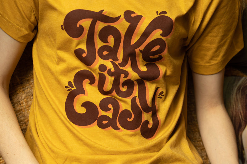 Smiling person wearing mustard yellow ‘Take It Easy’ graphic tee by Funnel Cake Tree with bold retro brown lettering, vintage style positive vibe t-shirt for laid-back days.