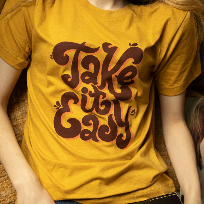 Smiling person wearing mustard yellow ‘Take It Easy’ graphic tee by Funnel Cake Tree with bold retro brown lettering, vintage style positive vibe t-shirt for laid-back days.