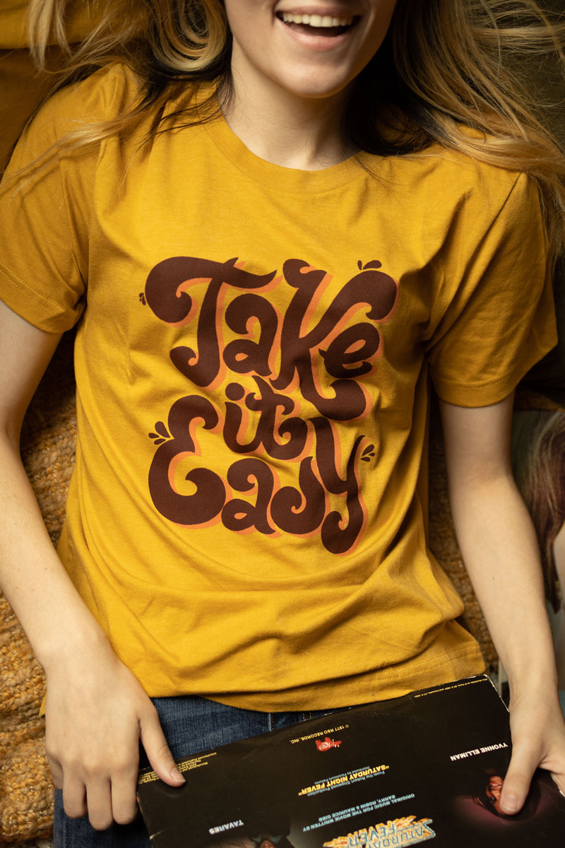 Smiling person wearing mustard yellow ‘Take It Easy’ graphic tee by Funnel Cake Tree with bold retro brown lettering, vintage style positive vibe t-shirt for laid-back days.