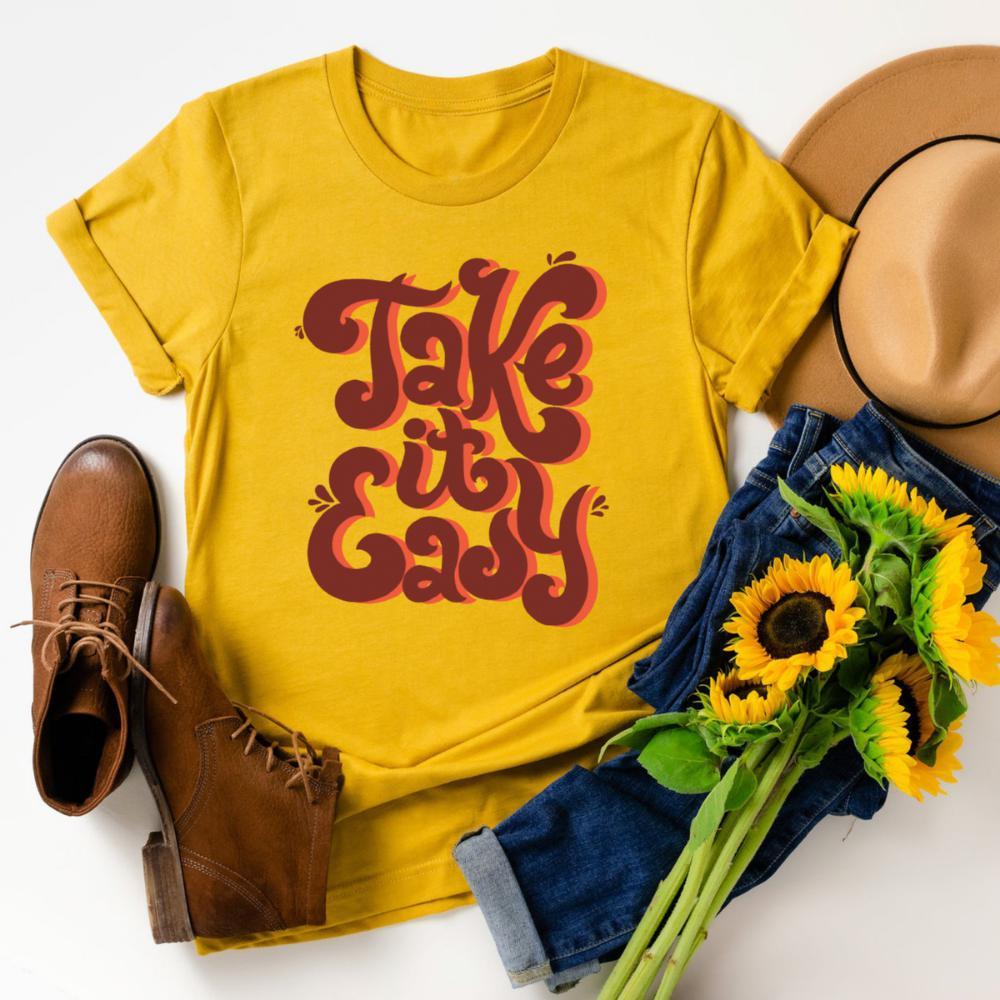 Flat lay of mustard yellow ‘Take It Easy’ graphic tee by Funnel Cake Tree with bold retro lettering in brown, styled with jeans, boots, sunflowers, and a hat, vintage positive vibes shirt.