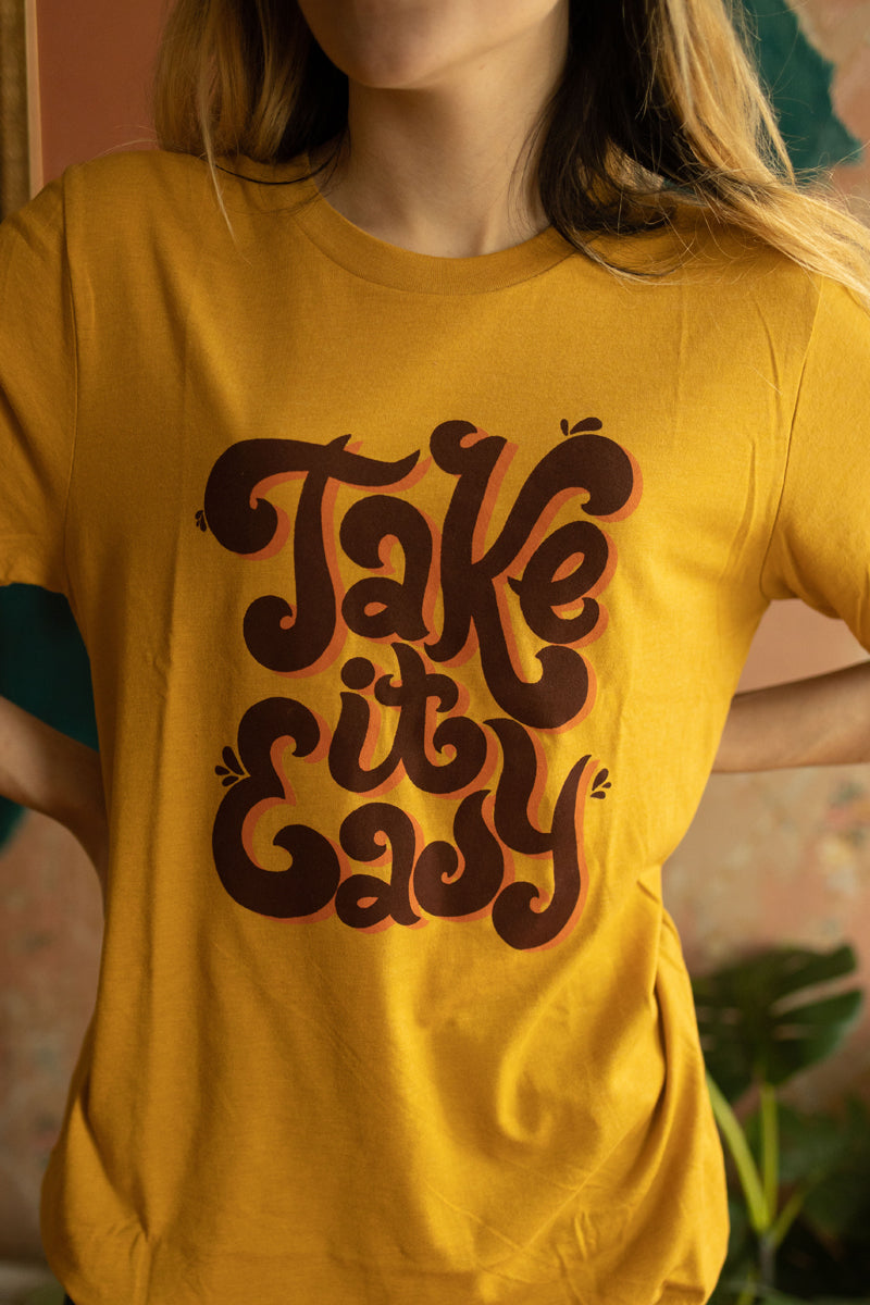 Close-up of mustard yellow ‘Take It Easy’ graphic tee by Funnel Cake Tree featuring bold retro brown lettering, vintage relaxed t-shirt with a groovy laid-back vibe.
