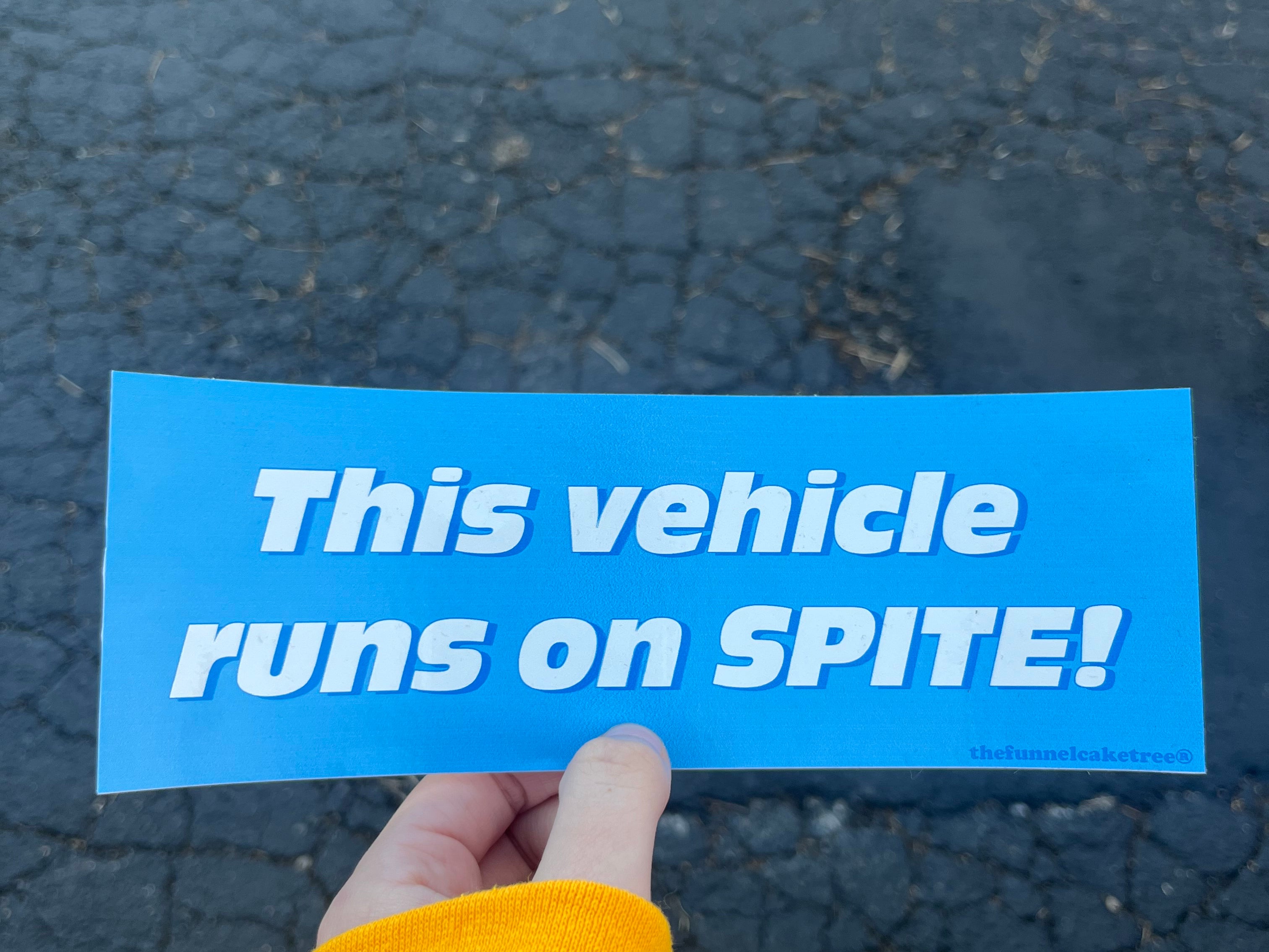 Glossy bright blue bumper sticker with bold white and blue text reading “This vehicle runs on spite!”