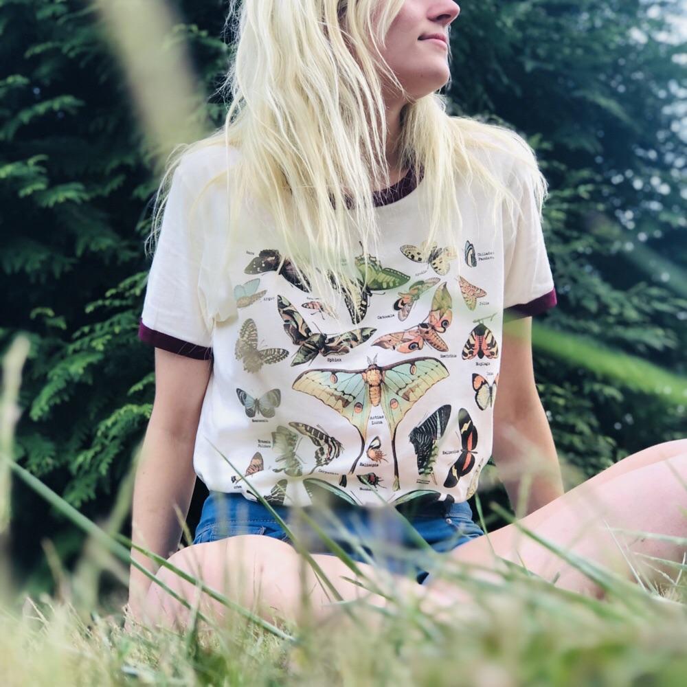 Woman sitting on grass wearing a vintage-style cream t-shirt with illustrated butterflies and moths, surrounded by greenery outdoors.