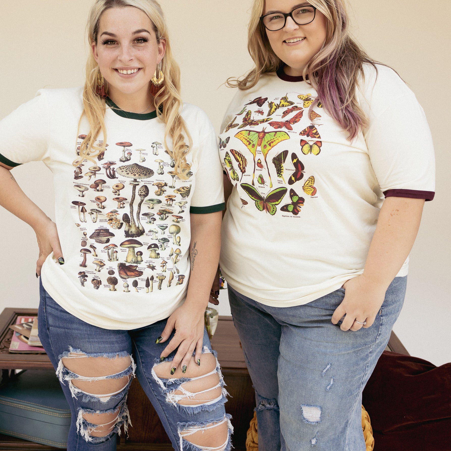 Two women wearing vintage-style nature graphic tees, one with illustrated mushrooms and the other with butterflies and moths standing together in casual jeans.