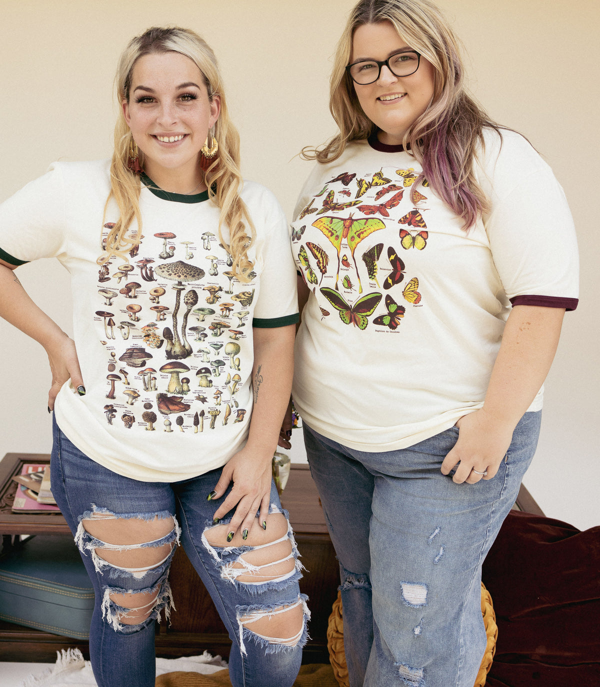 Two women wearing vintage-style nature graphic tees, one with illustrated mushrooms and the other with butterflies and moths standing together in casual jeans.
