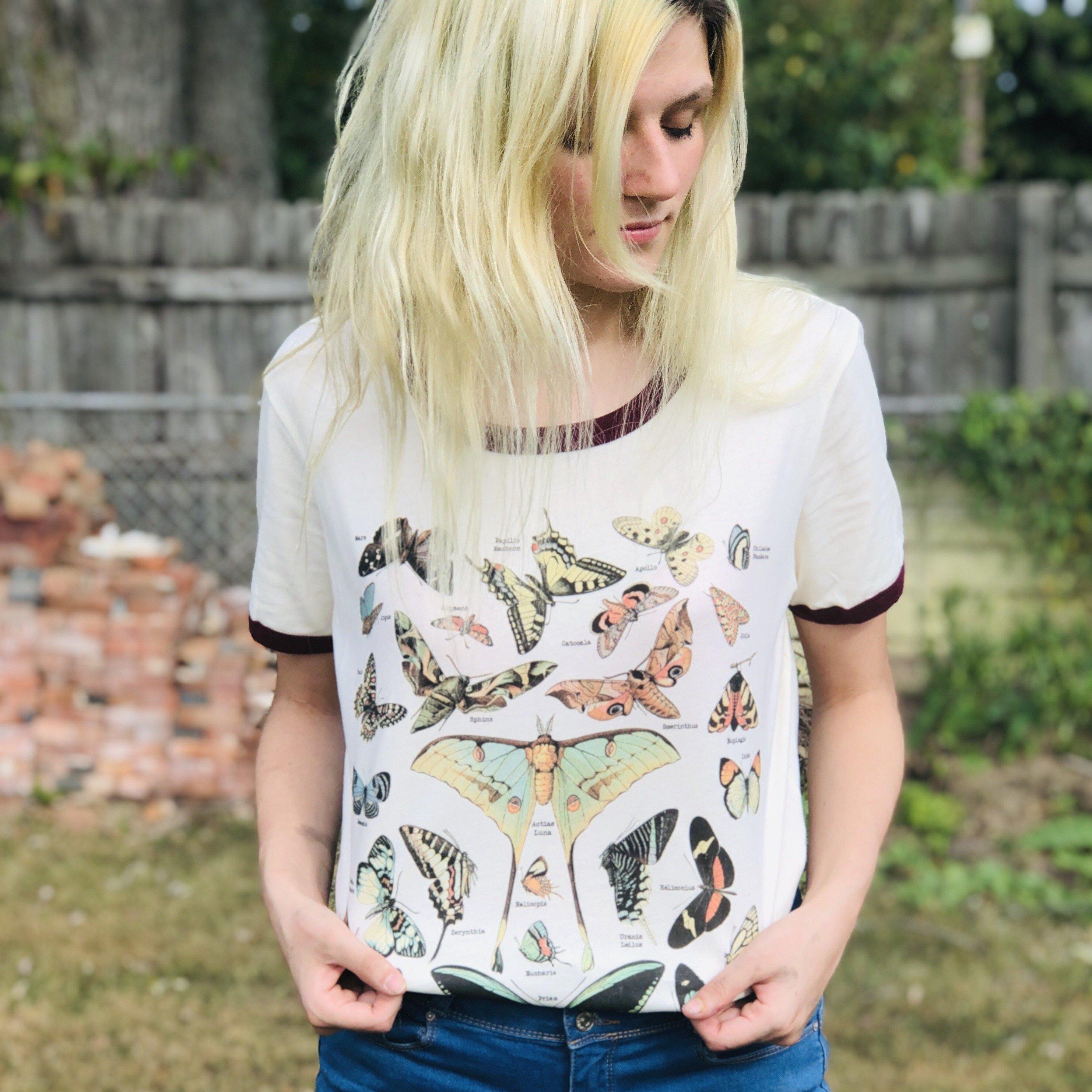Woman wearing a vintage-style cream t-shirt featuring illustrated butterflies and moths with scientific names, standing outdoors near a wooden fence.
