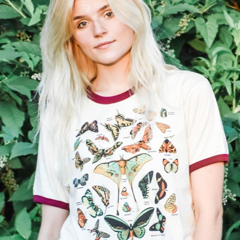Woman wearing a vintage-style ringer t-shirt with colorful illustrated butterflies and moths, standing outdoors in front of green leaves.