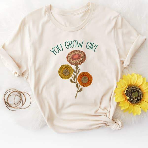 Flat lay of a cream ‘You Grow Girl’ graphic tee by Funnel Cake Tree featuring colorful retro floral design and sunflower accent, vintage botanical shirt celebrating growth and positivity.